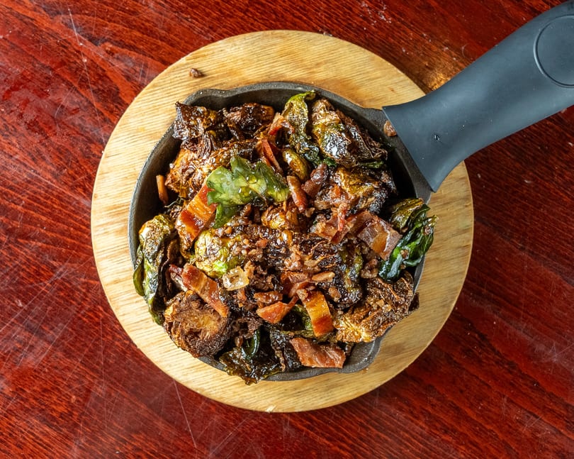Crispy Brussels sprouts with bacon bits served in a cast iron skillet on a wooden board.