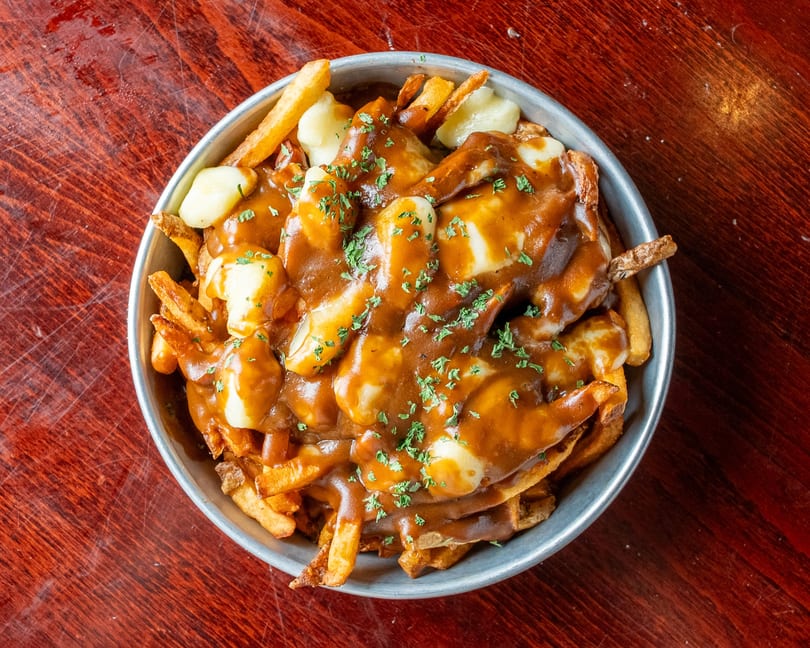 A metal bowl filled with golden french fries, cheese curds, and rich brown gravy, garnished with parsley.