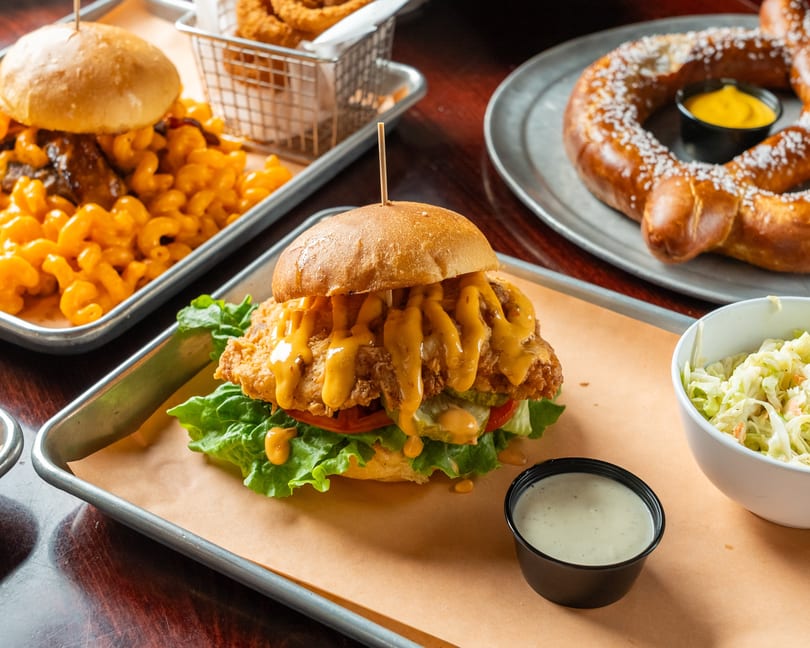 A fried chicken sandwich with cheese sauce, coleslaw, mac and cheese, and a pretzel with mustard.