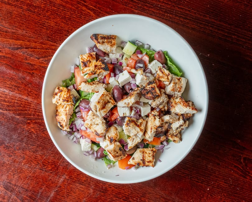 Overhead view of a grilled chicken salad with feta, olives, tomatoes, and red onion.