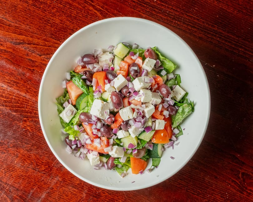 A vibrant Greek salad in a white bowl, featuring lettuce, tomatoes, cucumbers, olives, feta, and red onions.